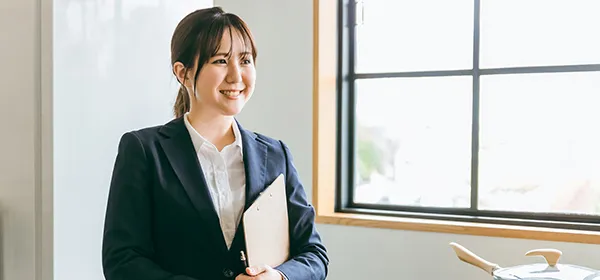バインダーを抱えて窓際に微笑み立っている女性社員の写真